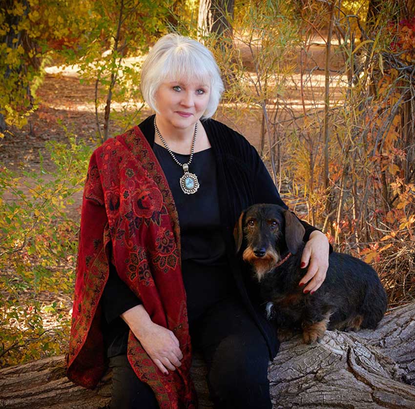 Woman with silver hair dressed in black with a red print scarf sitting on a log in an autumn background with her arm around a dachshund dog.